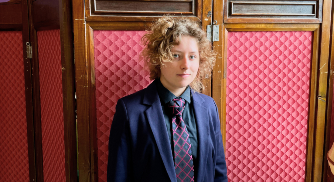 Kain, eine Person mit lockigen, dunkelblonden Haaren im Anzug.